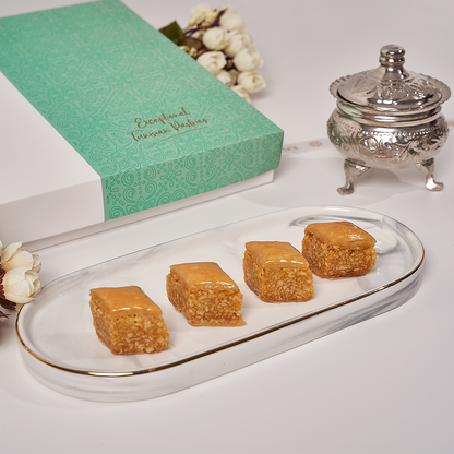 Side view of almond baklava pastries in a square gift box, featuring crispy filo layers and individually wrapped Mediterranean sweets