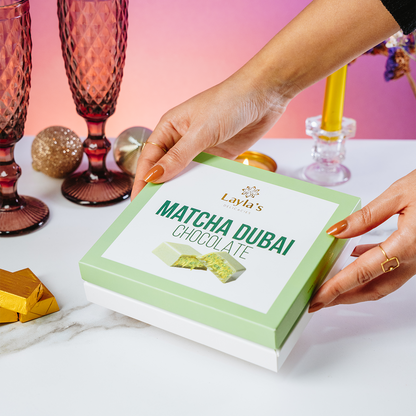 Hands holding a box of Layla's Matcha Dubai Chocolate with decorative items in the background.